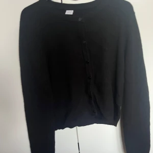 Svart kofta från Lindex med knappar - Svart, klassisk kofta från Lindex i storlek 170. Den har rund hals, långa ärmar och svarta knappar framtill. Materialet är mjukt och stickat, perfekt för att hålla värmen under kyliga dagar. Enkel och stilren design som funkar till många olika outfits.