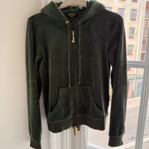 Mörkgrön velour hoodie Juicy Couture XS - Mörkgrön hoodie från Juicy Couture i mjuk velour med dragkedja och huva. Tröjan har långa ärmar, känguruficka och guldfärgad dragkedja med glittrande J-detalj. Perfekt för dig som gillar klassisk Y2K-stil och vill ha något riktigt cozy.
