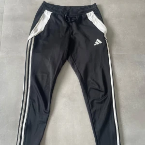 Svarta Adidas mjukisbyxor med vita detaljer - Svarta mjukisbyxor från Adidas med vita paneler vid fickorna och vita ränder längs benen. Byxorna har snörning i midjan och smal passform. Tillverkade i polyester, perfekt för träning eller chill. Adidas-logga på vänster lår för extra sportig vibe.