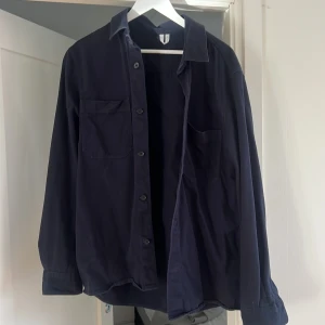 Mörkblå overshirt med fickor - Snygg mörkblå overshirt med klassisk krage och två bröstfickor. Modellen är rak och har knäppning framtill med svarta knappar. Perfekt att bära öppen över en t-shirt eller stängd som skjorta. Materialet känns slitstarkt och passar till många olika stilar.