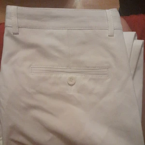 Vita kostymbyxor i polyester, stl 34 - Snygga vita kostymbyxor i 100% polyester med klassisk passform. Byxorna har bälteshällor, bakficka med knapp och stilren design. Perfekta för dig som vill ha en clean och fräsch look.