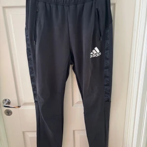 Svarta Adidas mjukisbyxor med logga - Svarta mjukisbyxor från Adidas med vit logga på låret och diskreta mönstrade sidopaneler. Byxorna har dragkedjefickor och elastisk midja. Perfekta för chill eller träning, och sportig passform.