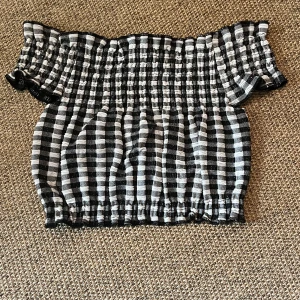 Svartvit rutig offshoulder topp - Säljer en svartvit rutig offshoulder topp med smockad och elastisk passform. Toppen har korta ärmar och är croppad, vilket ger en trendig vibe. Materialet är mjukt och stretchigt, perfekt för att styla med höga jeans eller kjol.