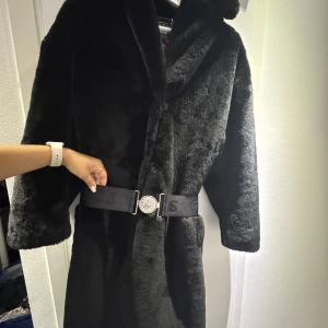 Svart pälsjacka från Guess med bälte - Snygg svart pälsjacka från Guess med fluffig känsla och markerad krage. Jackan har ett brett elastiskt bälte med Guess-logga och en cool silverfärgad spänndetalj. Perfekt för dig som vill sticka ut med en lyxig vibe under kalla dagar.