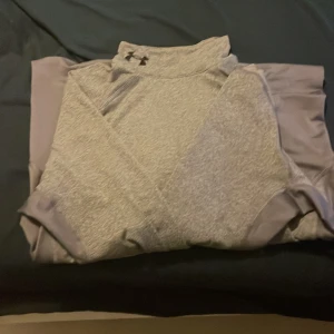 Grå långärmad träningströja Under Armour - Snygg grå långärmad träningströja från Under Armour med halv polo och diskret logga på både fram- och baksida. Materialet är mjukt och syntetiskt, perfekt för sport eller gym. Tröjan har en slim passform och paneler i mörkare grått på ryggen för extra stil.