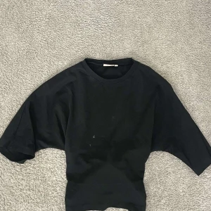 Svart croppad topp med knyt i ryggen - Svart croppad topp med vida ärmar och snygg knytning i ryggen. T-shirttyg i bomull gör den både bekväm och stilren. Perfekt för dig som gillar en enkel men cool look med en twist på ryggen.