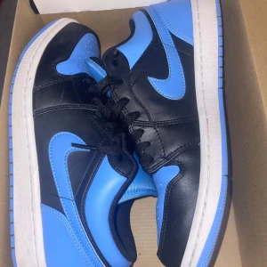 Nike Air Jordan 1 Low svart/blå - Säljer ett par Nike Air Jordan 1 Low i svart och ljusblått skinn med klassisk swoosh och Jumpman-logga på plösen. Skorna har svart snörning, perforerad tå och vit mellansula med blå yttersula. De har bara används två gånger men passar inte längre . Priset kan sänkas lite om det skulle behövas.
