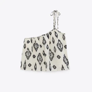 Vit och svart oneshoulder topp Zara - Säljer en luftig och cool oneshoulder topp med svart grafiskt mönster på vit bakgrund. Toppen har en smal axelrem med knyt och är croppad i modellen. Perfekt för varma dagar och ger en avslappnad vibe till din outfit.