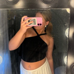 Svart croppad omlottopp med axelband - Säljer en svart croppad topp med snygg omlottdesign och korsade axelband framtill. Toppen är ärmlös och har en stilren look som passar perfekt till höga byxor eller kjol. Materialet känns mjukt och följsamt mot huden.