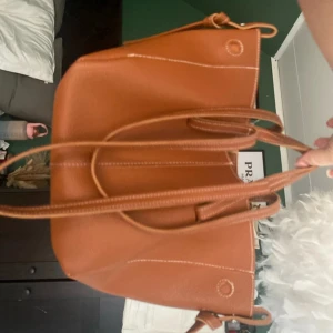 Brun axelväska i skinn med vita sömmar - Säljer en stilren brun axelväska i skinn med vita kontrastsömmar och dubbla handtag. Väskan har en rymlig design och är perfekt för dig som vill ha plats för allt du behöver under dagen. Enkel och snygg look som passar till många outfits. den har små skavanker i cirklarna på väskan som syns på bilderna men inget man märker alls, även en lös tråd men det påverkar inte funktionen då jag fått de fixat med textillim.