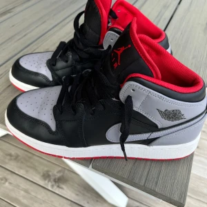Nike Air Jordan 1 Mid Svart/Röd/Grå - Nike Air Jordan 1 Mid i svart, grått och rött. Skorna har svart snörning, röd insida och vit sula med röd undersida. Tvättade. Använt sparsamt. Storlek junior 39 (24,5cm)  kommer med original box!