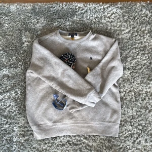 Grå sweatshirt från Polo Ralph Lauren - Snygg grå sweatshirt från Polo Ralph Lauren med klassisk rund hals och broderad marinblå logga på bröstet. Tröjan har långärmad passform och är gjord i mjuk bomullsmix som är skön mot huden. Perfekt för chill dagar och enkel att matcha med jeans eller joggers. Passar killar som har S