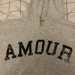 Grå hoodie med svart tryck och stjärna där bak  - Snygg ljusgrå hoodie med svart text 'AMOUR' framtill och en cool svart stjärna på ryggen. Tröjan har huva och är tillverkad i mjukt material, perfekt för chill dagar. Passar dig som gillar enkel men trendig stil.