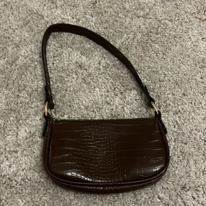 Brun axelväska från asos - Snygg brun axelväska i speciellt mönster och guldfärgade detaljer. Väskan har en stilren, avlång form och stängs med dragkedja. Perfekt storlek för det viktigaste. Materialet är glansigt och ger en lyxig känsla. Ny pris 319kr