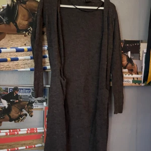 Mörkbrun lång stickad kofta  Vila - Lång, mörkbrun stickad kofta från Vila med lång ärm och enkel design. Koftan har två fickor framtill och är perfekt för dig som gillar en chill och stilren look. Mjuk och skön, passar till många olika outfits.