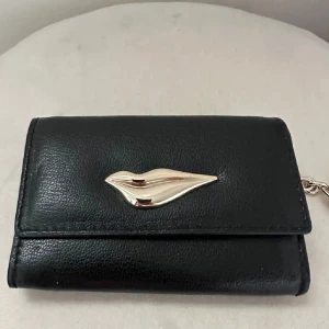 Svart plånbok från Diane von Furstenberg - Svart plånbok i från DVF med guldfärgad läppdetalj på framsidan och nyckelring i metall. Insidan har svartvitt och ett fack. Perfekt för kort, mynt och nycklar
