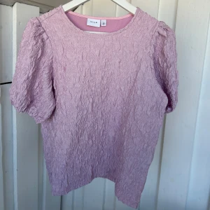 Ljusrosa blus från Vila, S - Säljer en ljusrosa blus från Vila i storlek S. Blusen har korta puffärmar och ett coolt, krinklat tyg som ger en unik struktur. Rund halsringning och loose passform gör den både bekväm och trendig. Perfekt för dig som gillar att sticka ut med färg och textur.