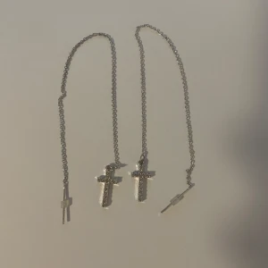 Silvriga örhängen med kors och kedja - Säljer ett par långa silvriga örhängen med tunn kedja och glittrande kors i änden. Korsen är täckta av små stenar som ger extra bling. Perfekt för dig som gillar statement-smycken med lite edge. Kedjan är ca 17 cm lång och träs igenom örat. Små missfärgningar på kedjan om ses på andra bilden.
