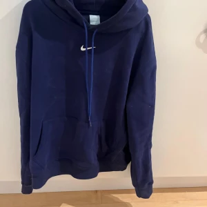 Mörkblå Nike NOCTA hoodie med huva - Säljer en mörkblå hoodie från Nike NOCTA med klassisk huva och snörning. Tröjan har en gul Nike-logga på bröstet, känguruficka framtill och A.W.R.-brodyr på ryggen. Materialet är mjuk bomull, perfekt för chill dagar. Passformen är loose och ärmarna är långärmade.