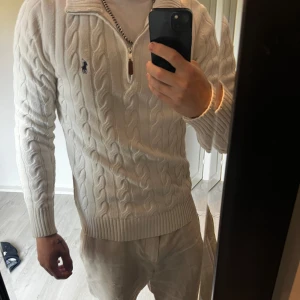 Halv zip från Polo Ralph Lauren - Säljer en vit kabelstickad tröja från Polo Ralph Lauren. Tröjan är original XL men minskad i högvärme tvätt, inget som syns på själva designen och är som en Large nu. Klassisk broderad logga på bröstet och riktigt skön passform. Perfekt för dig som gillar stilren och fräsch design.