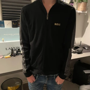 Svart zipjacka från BOSS Homewear - Svart zipjacka från BOSS Homewear med gråa paneler längs ärmarna och guldfärgad BOSS-logga på bröstet. Jackan har hög krage, hel dragkedja och ribbade muddar. Perfekt för en chill och stilren look. Storlek S