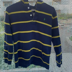 Randig marinblå hoodie från Polo Ralph Lauren - Säljer en marinblå hoodie från Polo Ralph Lauren med gula ränder och grönt broderat logotyp på bröstet. Tröjan har huva och långa ärmar. Materialet är bomull och polyester, vilket gör den både mjuk och skön. Perfekt för chill dagar och för en mer vågad stil.