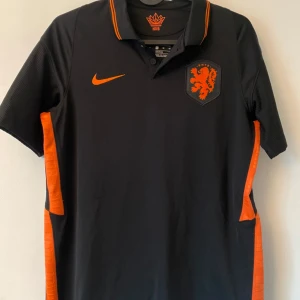 Holländsk Nike fotbollströja  - Snygg svart fotbollströja från Nike med orange detaljer och KNVB-logga på bröstet. Tröjan har korta ärmar, orange Nike-märke och ränder längs sidorna. Tillverkad i Dri-FIT-material som andas, perfekt för träning eller match.