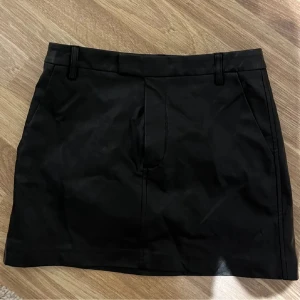Svart skinnkjol - Snygg svart kortkjol med klassisk rak passform och praktiska fickor framtill. Kjolen har hällor för bälte och stilrena sömmar som ger en clean look. Perfekt för dig som gillar minimalistisk stil och vill ha en enkel kjol som funkar till många outfits.