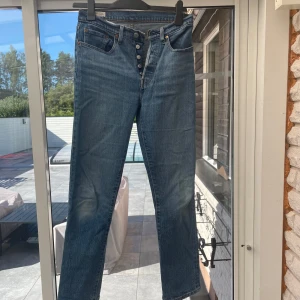 Levi's 501 blå jeans straight fit - Klassiska Levi's 501 jeans i blå denim med raka ben och knäppning framtill. Jeansen har fem fickor, normal passform och ikonisk röd Levi's-tag bak. Perfekta för en avslappnad och tidlös stil.
