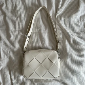 Vit quiltad axelväska i skinnimitation - Snygg vit axelväska med quiltad design och bred axelrem. Väskan har silverfärgade detaljer och dragkedja upptill. Perfekt storlek för det viktigaste och ger en clean look till din outfit.