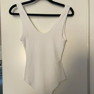 Vit ärmlös bodysuit med djup rygg - Säljer en stilren vit bodysuit med djup urringning både fram och bak. Modellen är ärmlös och har breda axelband. Perfekt att styla med jeans eller kjol för en clean look. Mjuk och stretchig i materialet för en bekväm passform.