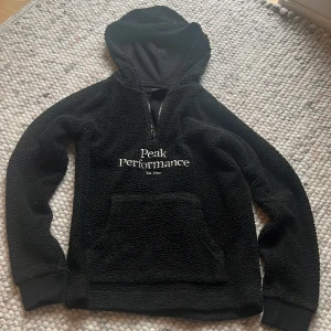 Svart fleecehoodie Peak Performance - Mysig svart hoodie från Peak Performance i fleece med broderad logga på bröstet. Tröjan har huva, halv dragkedja och stor känguruficka framtill. Perfekt för kyliga dagar och chill vibes.
