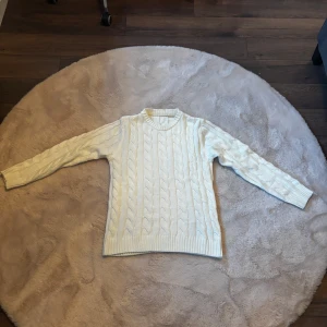Vit kabelstickad tröja med rund hals - Säljer en vit kabelstickad tröja med rund hals och långa ärmar. Tröjan har ett klassiskt flätmönster och ribbade muddar vid hals, ärmslut och nederkant. Perfekt för dig som gillar en clean och stilren look under höst och vinter.