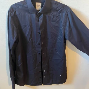 Svart skjorta Oscar of Sweden - Snygg svart skjorta från Oscar of Sweden i slim fit-modell. Skjortan har klassisk krage, svarta knappar och lång ärm. Materialet är bomull och är wrinkle free, vilket gör den enkel att hålla fräsch. Perfekt för dig som gillar stilrena och moderna plagg.