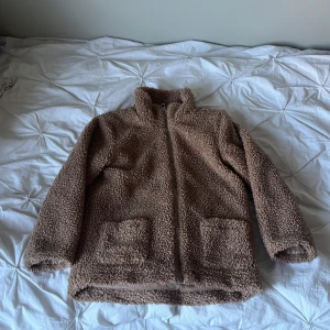Brun teddyjacka från KappAhl stl 128 - Mysig brun teddyjacka från KappAhl i storlek 128. Jackan har två stora fickor framtill, dragkedja och hög krage. Perfekt för kyliga dagar och ger en riktigt skön vibe med sitt fluffiga material.