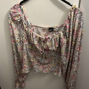 Blommig blus med knyt från Gina Tricot - Supersöt blommig blus från Gina Tricot i tunnt, luftigt material. Toppen har långa ärmar med resår vid handlederna, fyrkantig ringning och knyt fram. Färgerna går i lila, rosa, gul och vit, perfekt för en fräsch look.