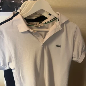 Vit pikétröja från Lacoste, S - Vit pikétröja från Lacoste med klassisk krage, knäppning framtill och den ikoniska krokodilloggan på bröstet. Tröjan har gröna och svarta detaljer på insidan av kragen och är tillverkad i bomull. Perfekt för en clean och sportig stil.
