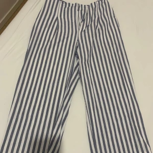 Randiga blåvita tygbyxor - Snygga linnebyxor med vertikala blå och vita ränder. Byxorna har en rak och loose passform med hög midja och är tillverkade i 100% bomull. Perfekta för dig som gillar en chill och stilren look med lite marint vibe.