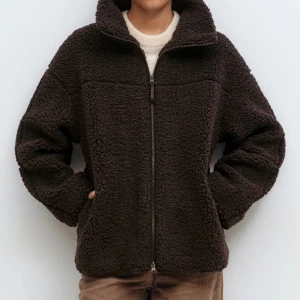 Mörkbrun teddyjacka med dragkedja - Mysig teddyjacka i mörkbrunt med hög krage och hel dragkedja framtill. Jackan har en loose passform och är tillverkad i mjukt fleece-material som håller dig varm. Perfekt för kyliga dagar och enkel att matcha med dina favoritjeans eller byxor.