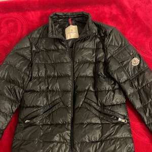 Svart Moncler dunjacka med dragkedja - Säljer en svart dunjacka från Moncler med klassisk quiltad design och dragkedja framtill. Jackan har två snedställda fickor med dragkedja och Moncler-logga på ärmen. Perfekt för kyliga dagar och riktigt snygg till streetwear-stilen.