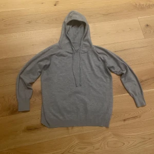 Grå merinoull hoodie med huva och snörning - En ALDRIG ANVÄND stilren grå hoodie med huva och snörning. Tröjan har lång ärm och ribbade muddar vid ärmslut och nederkant. Perfekt för dig som gillar en enkel och clean look. Materialet känns mjukt och passar bra till chill dagar.