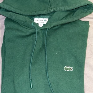 Grön hoodie från Lacoste Classic Fit - Mörkgrön hoodie från Lacoste i klassisk passform med broderad krokodil-logga på bröstet. Tröjan har huva med snörning, magficka och ribbade muddar. Perfekt för dig som gillar stilren look. Inte alls mycket använd på grund av att den var i fel köpt i för storlek. Priset kan diskuteras 💚