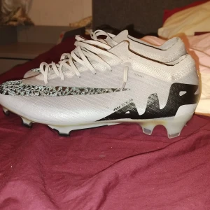 Nike Air Zoom fotbollsskor vita - Snygga vita Nike Air Zoom fotbollsskor med svart och glittrigt mönster på swoosh-loggan. Skorna har snörning, låg profil och en cool iriserande sula med lila detaljer. Ovandelen är i syntetmaterial och mesh för bra andningsförmåga.
