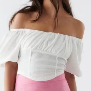 Vit offshoulder korsett-topp - Superfin vit korsett-topp med offshoulder och puffiga korta ärmar. Toppen är croppad och har en lätt rynkad struktur framtill som ger en snygg form. Perfekt till höga jeans eller kjol för en trendig look.