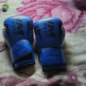 Blå boxningshandskar HUANMENG 12oz - Säljer ett par blå boxningshandskar från HUANMENG med svarta detaljer och tryck. Handskarna har bred kardborre runt handleden för extra stöd och är designade för träning. Snygg sportig look med svart logga och text på ovansidan.