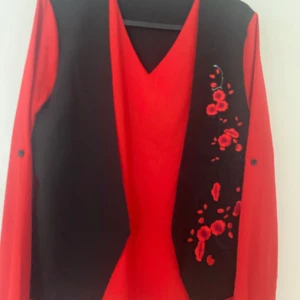 Svart väst med broderade röda blommor - Svart väst med v-ringad öppning och broderade röda blommor på ena sidan. Västen har en klassisk rak passform och är i ett mjukt tyg, perfekt att slänga över en skjorta eller topp för en cool look.