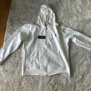 Vit hoodie från Polo Ralph Lauren - Snygg vit hoodie från Polo Ralph Lauren med dragkedja och klassisk broderad logga på bröstet. Tröjan har huva med snörning och två fickor framtill. Perfekt för en clean och stilren look. Storlek m men passar L