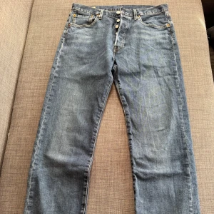 Levi's 501 blå jeans W31 L34 - Klassiska Levi's 501 jeans i blå denim med raka ben och knappgylf. Snyggt slitna detaljer och ikonisk läderpatch bak i midjan. Jeansen har fem fickor och orange sömmar. Perfekta för dig som gillar tidlös stil och vill ha ett par äkta Levi's.