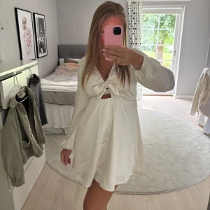 Vit kort klänning med rosätt - Supersöt vit klänning med lång ärm och knyt framtill som ger en snygg cut-out detalj. Klänningen har en flowy passform och är kort. Materialet är siden liknande. Säljer då den inte kommer till användning ❣️