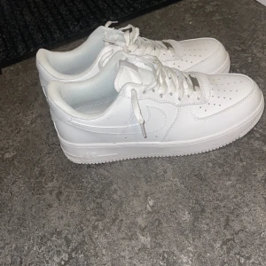 Nike Air Force 1 vita sneakers - Fräscha Nike Air Force 1 sneakers i helvitt skinn med klassisk låg siluett. Skorna har perforerad tå, snörning med metalltips och den ikoniska swoosh-loggan på sidorna. Yttersulan är platt och ger bra grepp. Perfekt för dig som gillar cleana och stilrena kicks.
